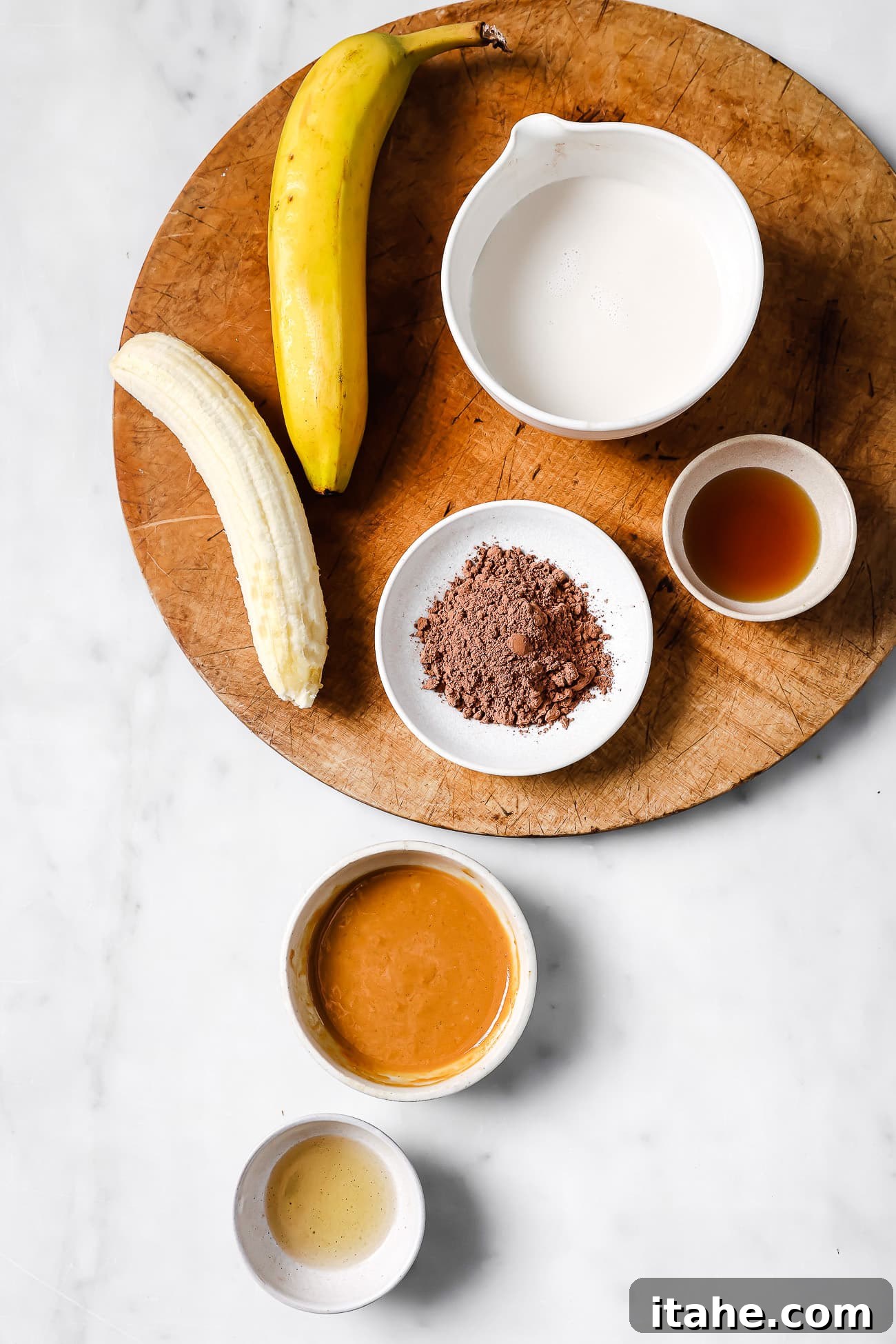 Pre-measured ingredients for a chocolate peanut butter smoothie laid out on a wooden cutting board and white countertop.