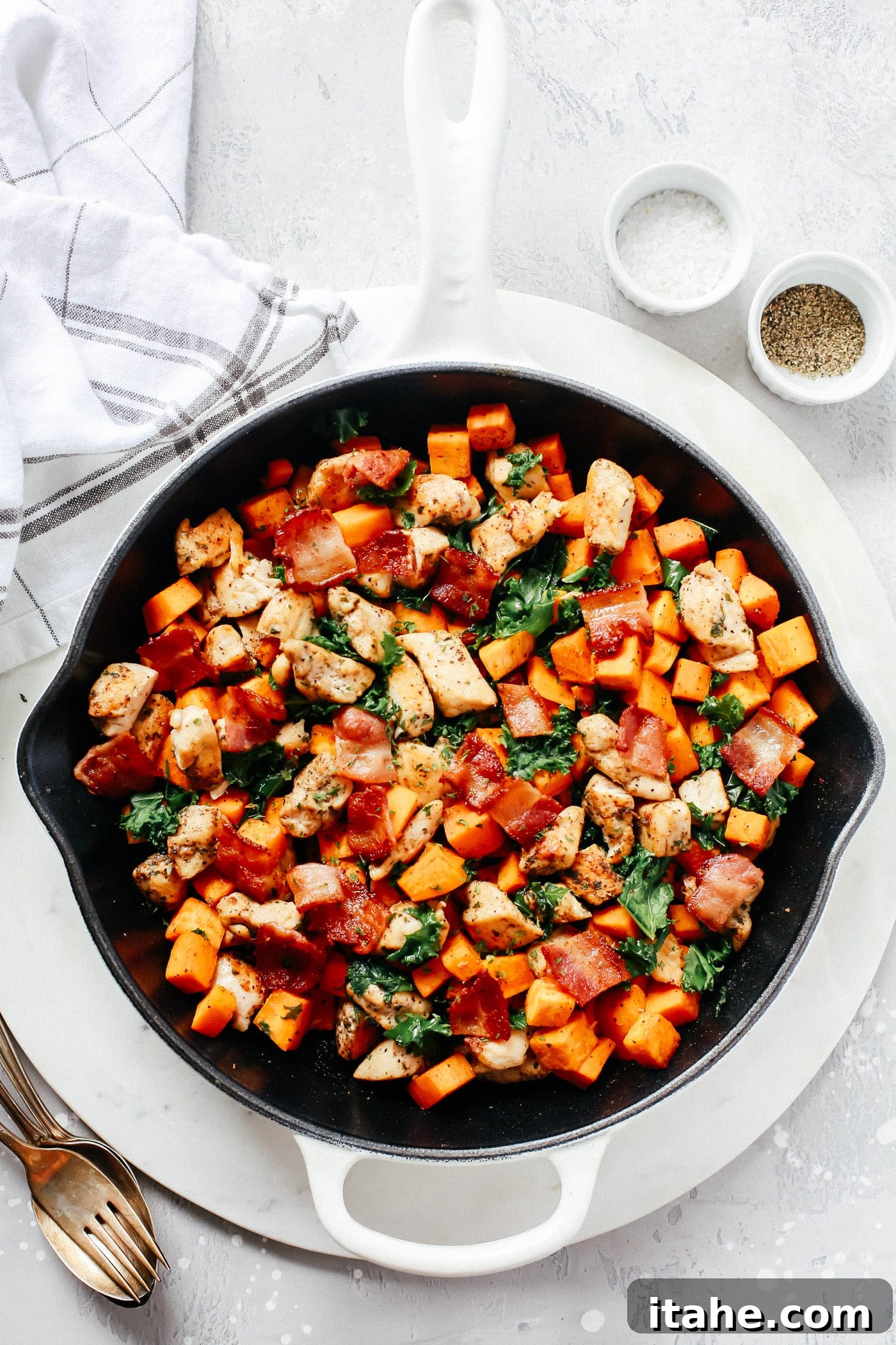 A cast iron skillet filled with diced chicken, sweet potato cubes, and vibrant green kale, topped with crispy bacon bits.