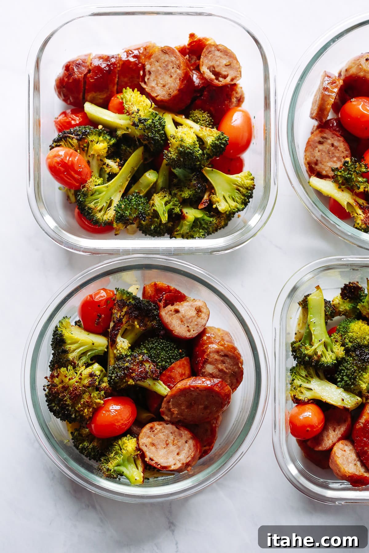Four glass bowls filled with savory Italian sausage, vibrant broccoli florets, and bright cherry tomatoes, perfectly portioned for meal prep, arranged on a rustic wooden surface.