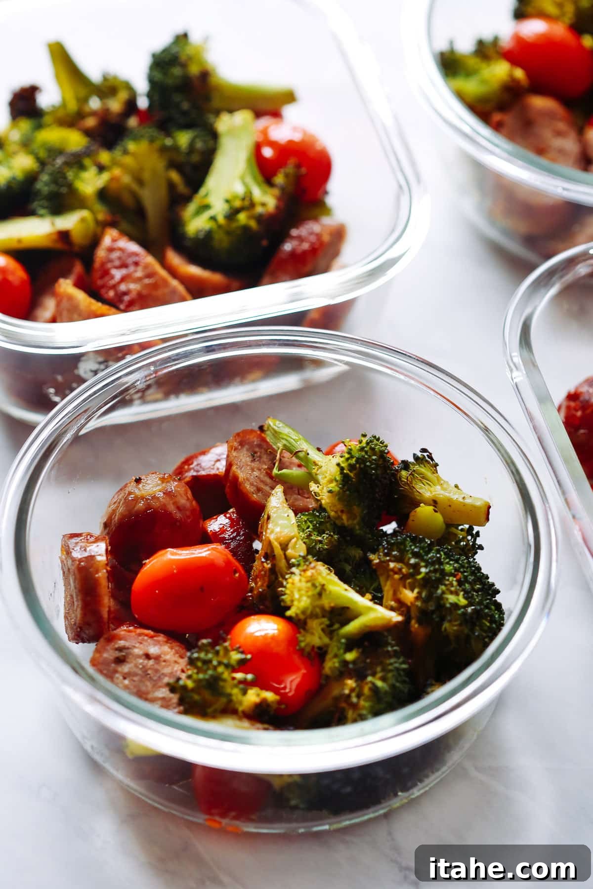 Close-up overhead shot of a single glass meal prep bowl featuring perfectly roasted Italian sausage, bright green broccoli florets, and juicy red cherry tomatoes, garnished with fresh herbs.