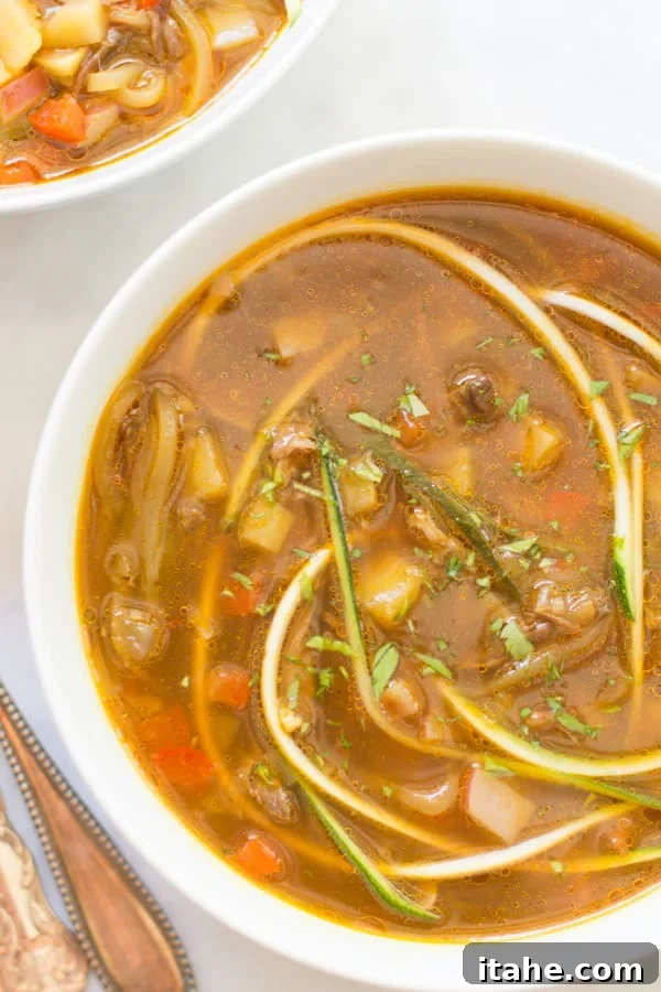 A close-up view of a steaming bowl of Zucchini Noodle Soup, garnished with fresh parsley.