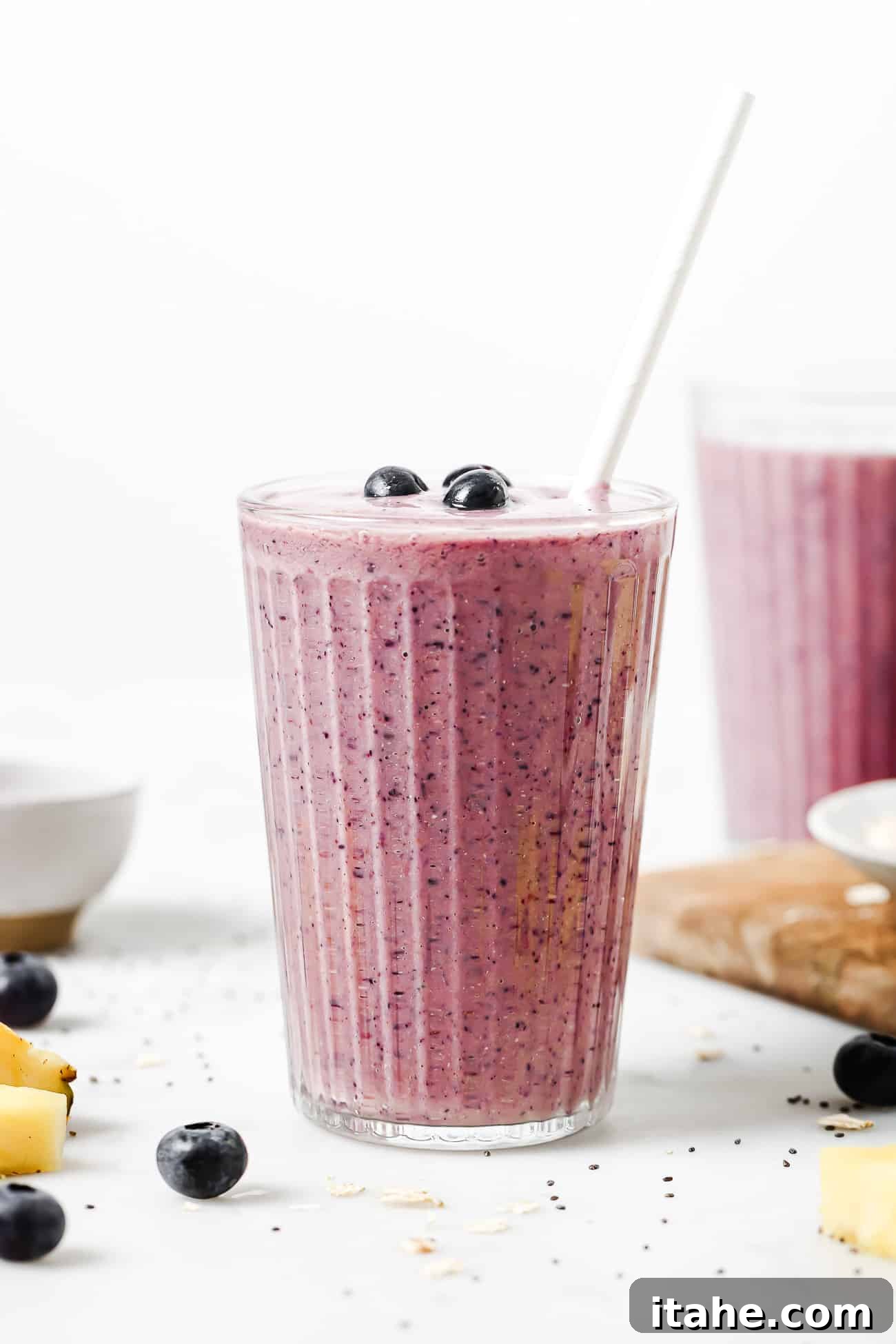 A vibrant, rich purple blueberry banana smoothie presented in a tall, slender glass with a straw, garnished with fresh blueberries, on a light background.