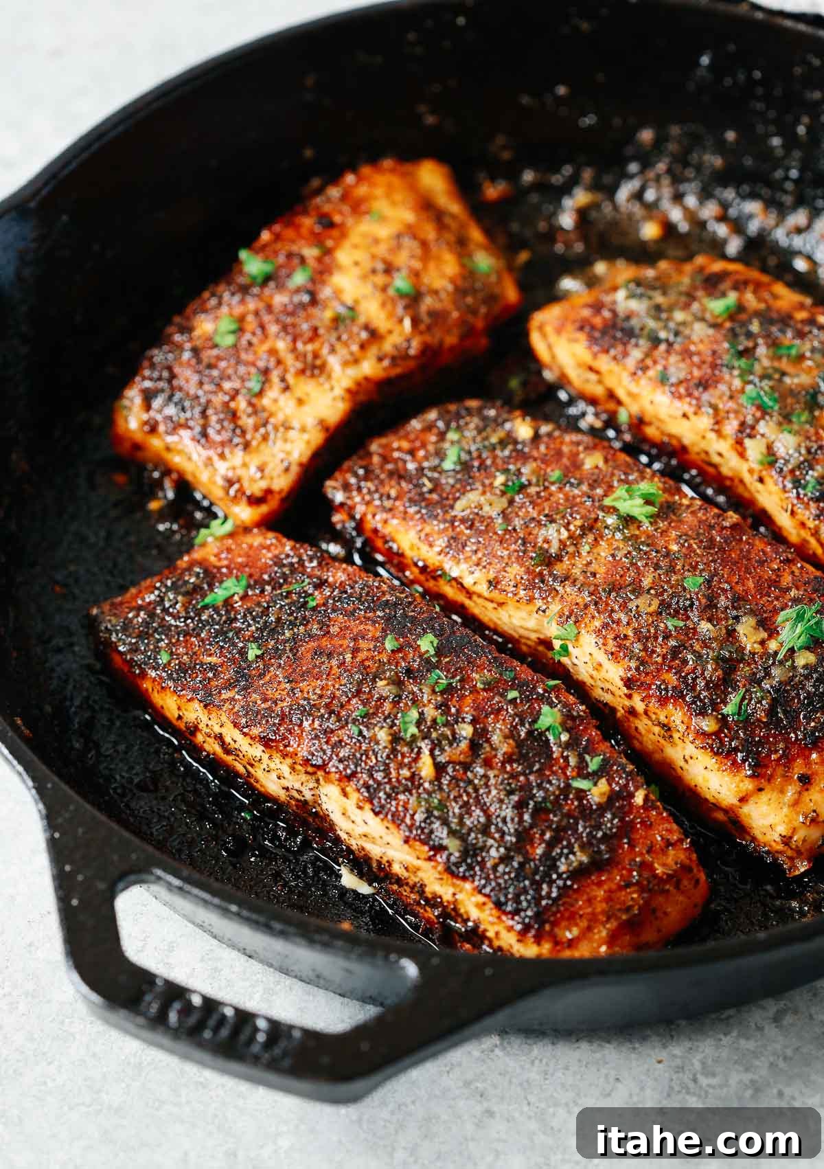Four perfectly blackened salmon fillets resting in a cast iron skillet, garnished with fresh herbs, ready to be served.