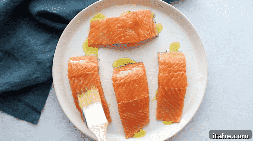 An overhead view of a salmon fillet being brushed with olive oil on a white plate.