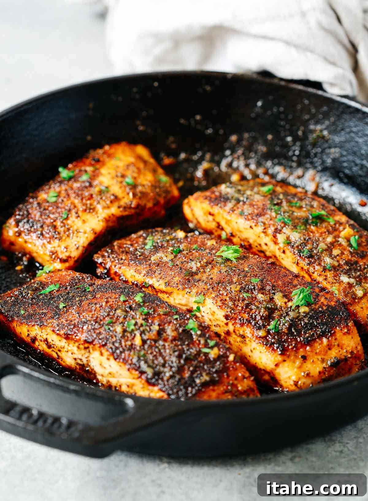 Four heavily seasoned salmon fillets being cooked in a hot cast iron skillet, showing the beginning stages of blackening.