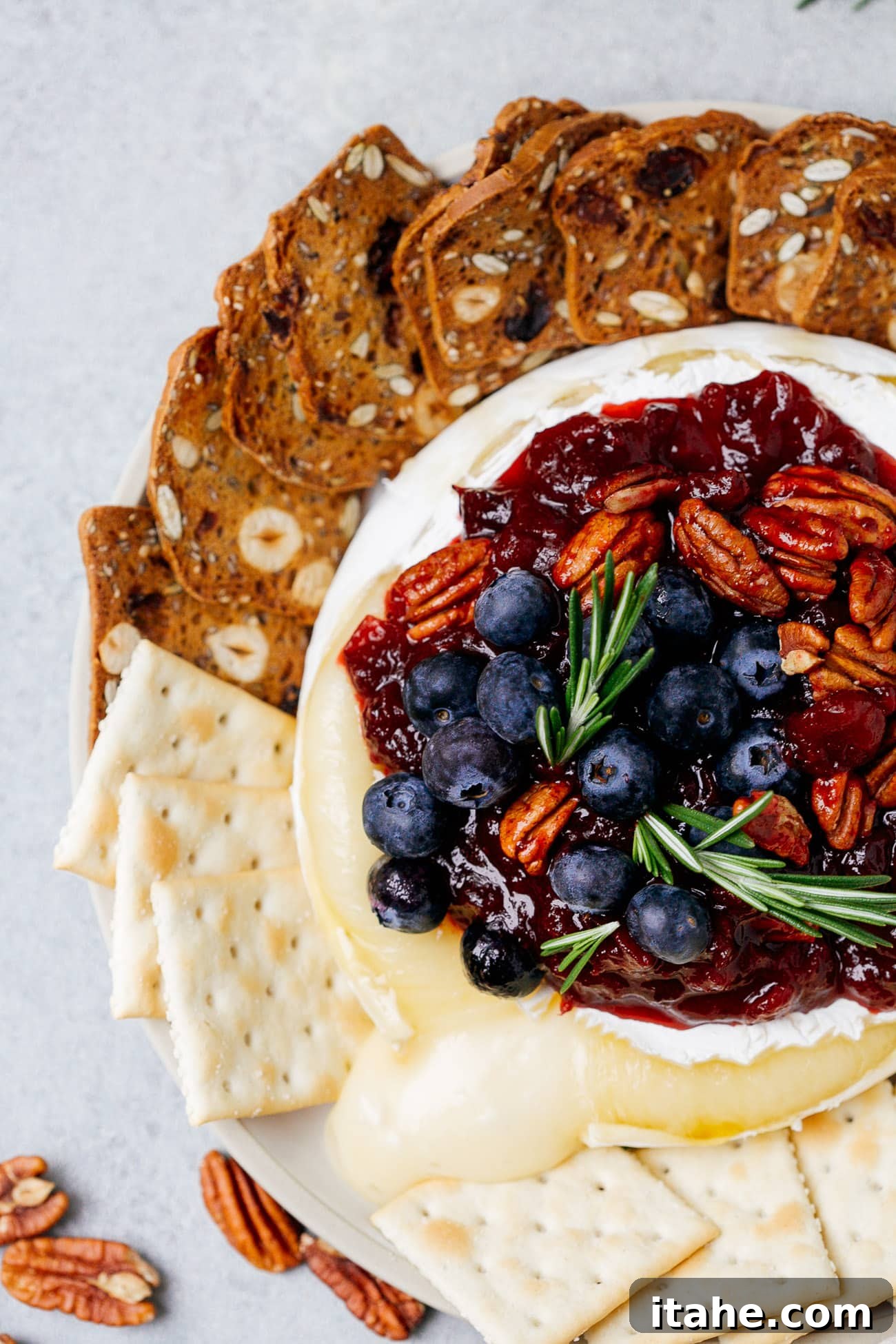 Warm Cranberry Brie Delight 4 Overhead view of a baked brie with cranberry sauce with pecans, blueberries, and rosemary on top, served with crackers.