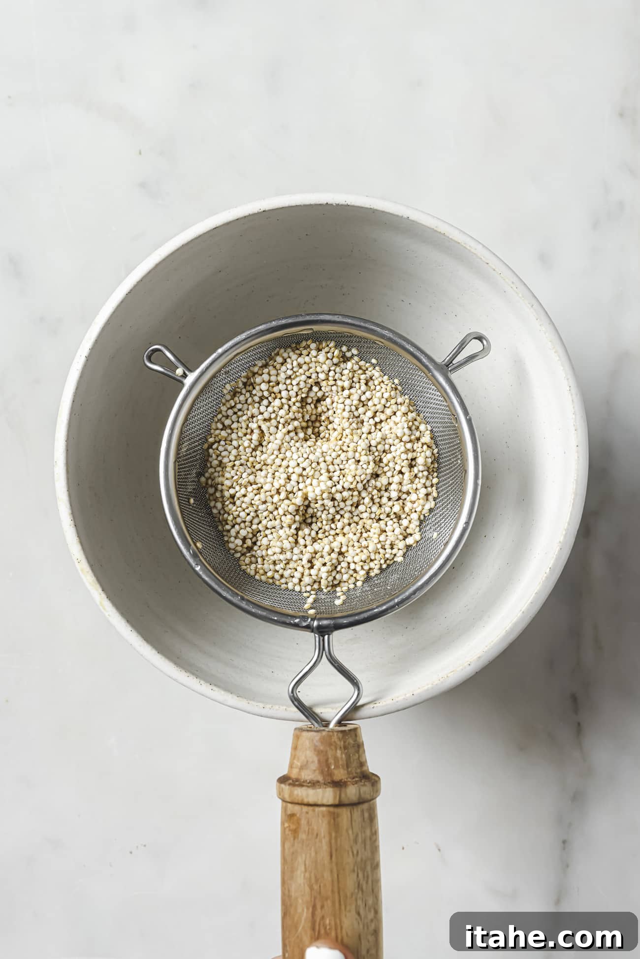 Uncooked quinoa in a fine mesh strainer over a white bowl. 