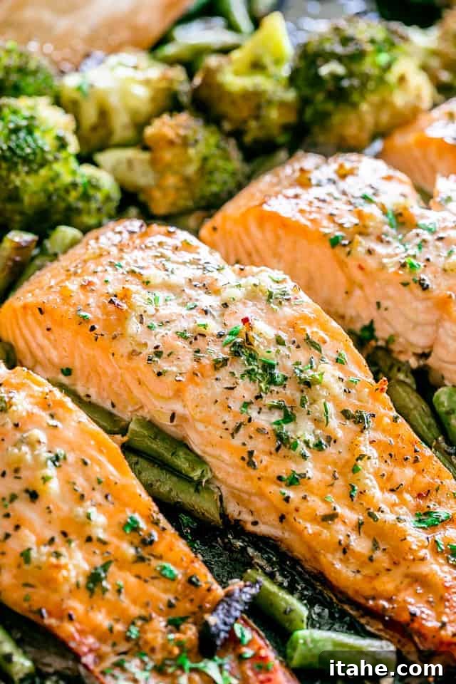 Three pieces of baked salmon on a baking sheet, accompanied by vibrant green beans and broccoli.