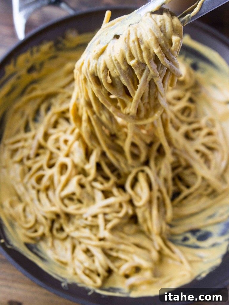 The Ultimate Fall Pumpkin Sauces 6 A pair of tongs delicately lifting a generous serving of fettuccine noodles, perfectly coated in a vibrant and creamy vegan pumpkin sauce.