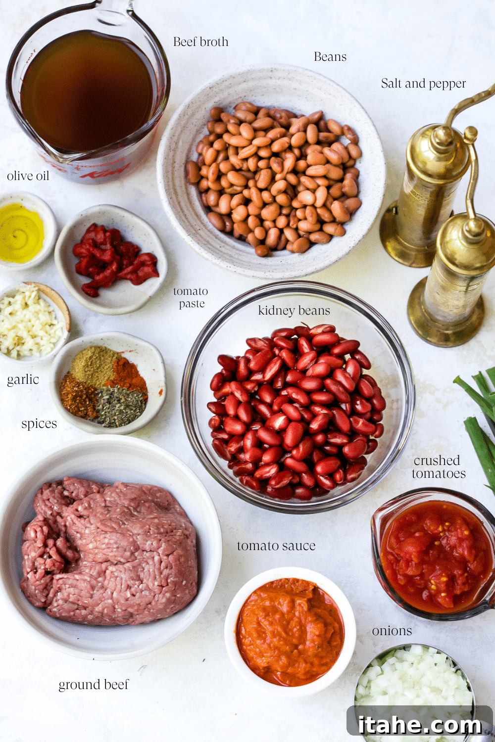 Ingredients laid out on a countertop, including olive oil, onions, garlic, ground beef, various tomato products, beans, broth, and spices, to make Classic Chili Recipe.
