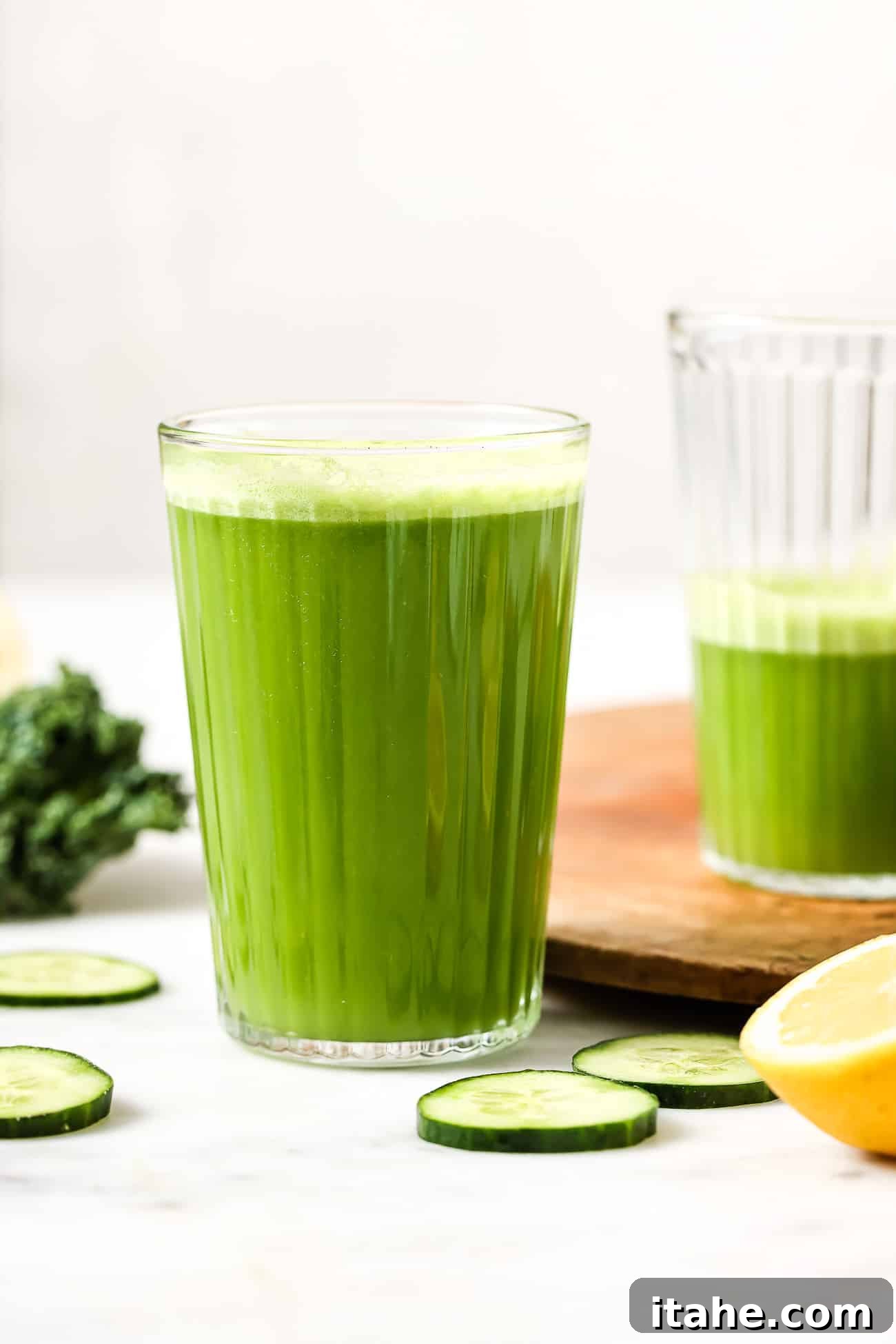 A glass of vibrant green juice, garnished with a slice of lemon, with fresh kale and cucumbers blurred in the background, suggesting natural ingredients.