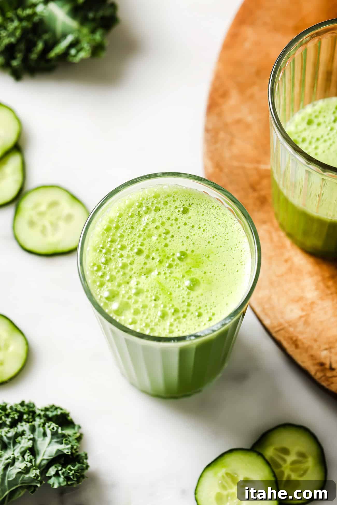 A close-up of a glass of green juice, featuring ice cubes, with fresh cucumbers and kale artfully arranged in the background, highlighting the natural ingredients.