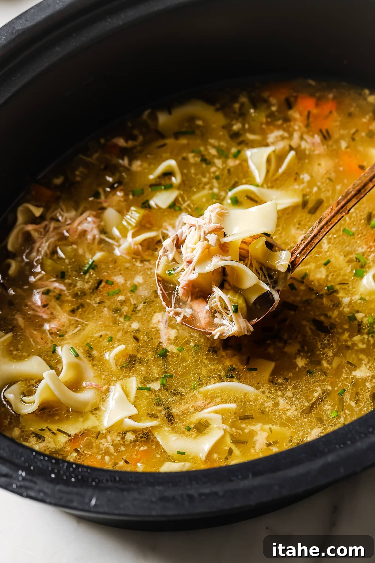 A close-up view of a bowl of slow cooker chicken noodles with a ladle of soup, garnished with fresh chives.