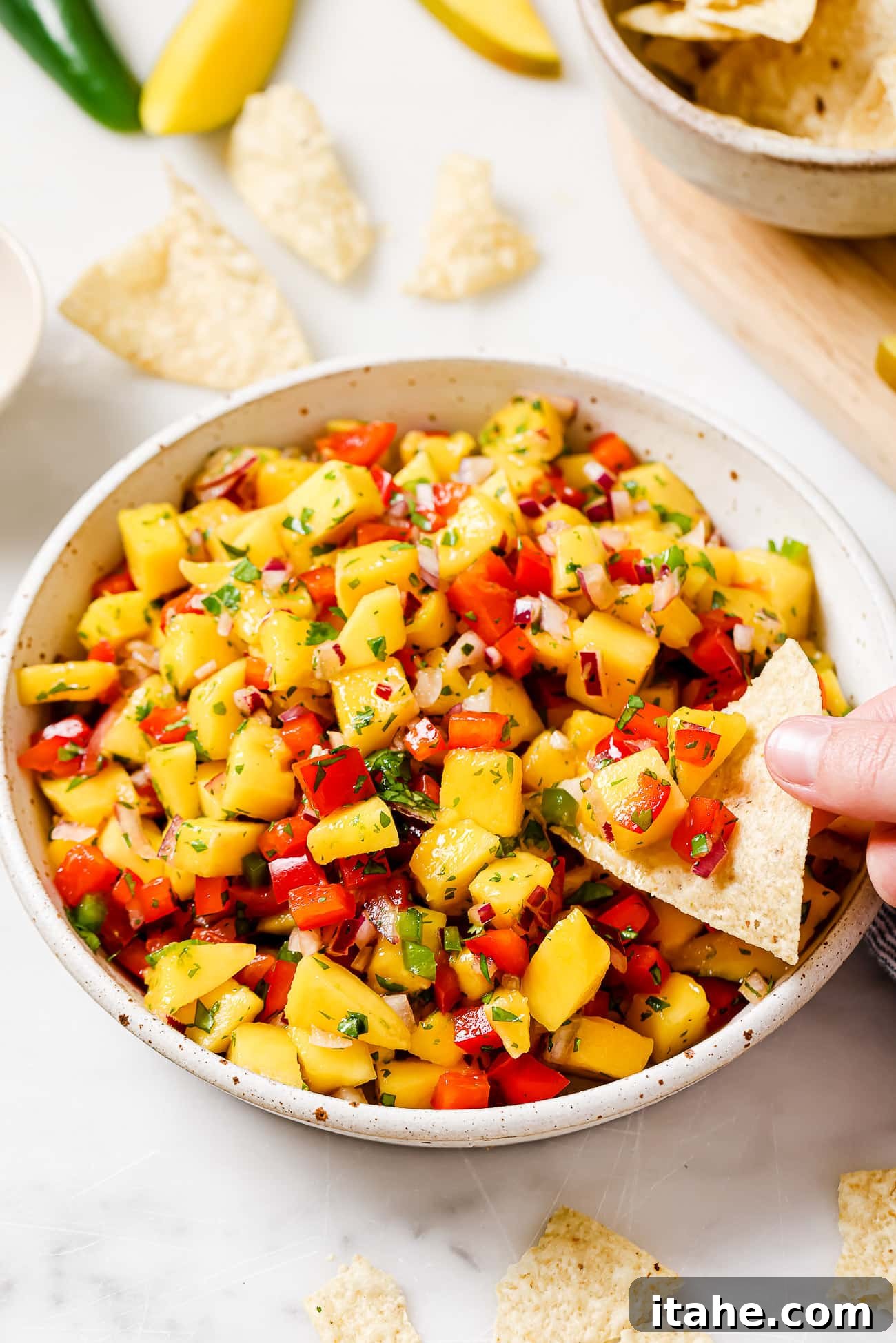 Tropical Mango Salsa 2 An overhead view of a tortilla chip scooping vibrant mango salsa from a large white bowl, showcasing the fresh diced ingredients.