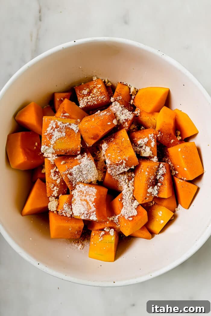 An overhead view of butternut squash and seasonings in a large mixing bowl, ready to be tossed.