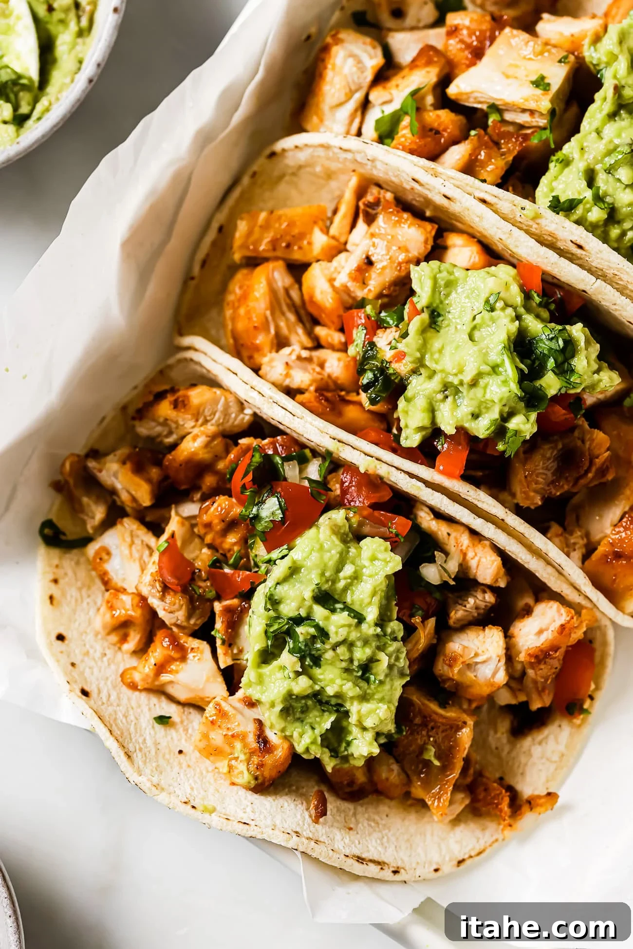 Three beautifully presented chicken tacos, richly topped with Pico de Gallo and creamy guacamole, resting elegantly on parchment paper, highlighting their appetizing appeal.