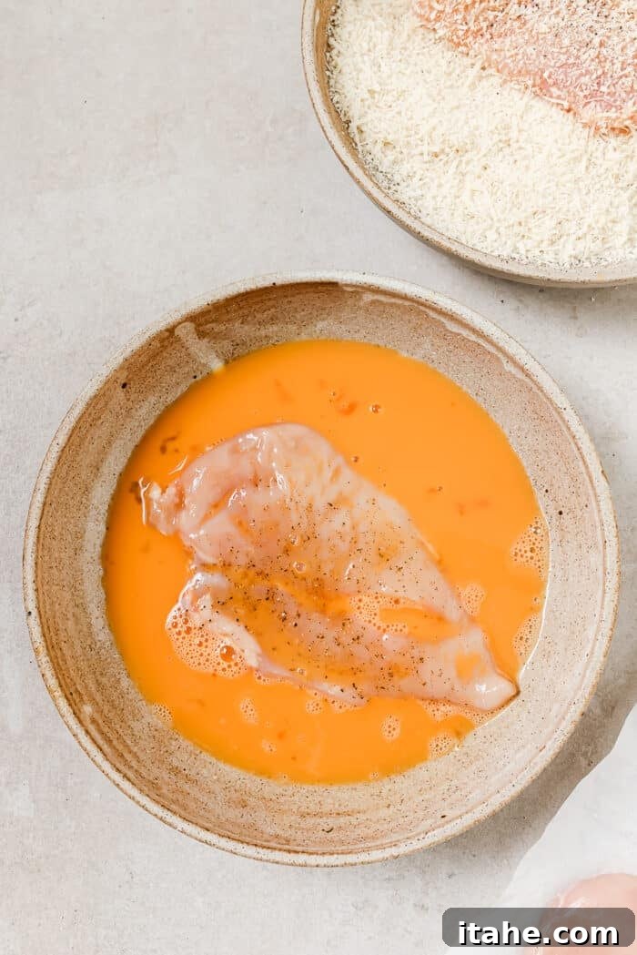 A chicken cutlet being dipped in a bowl of beaten eggs, with the breadcrumb mixture visible in the background.