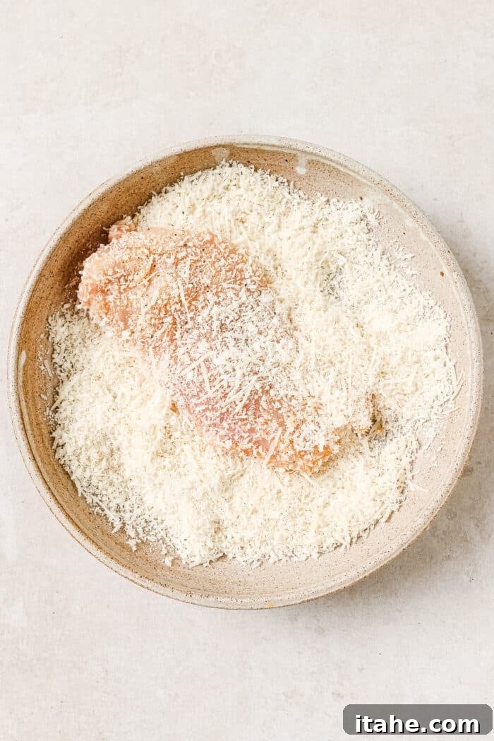 A chicken cutlet being pressed into a bowl of Panko breadcrumbs and Parmesan cheese.