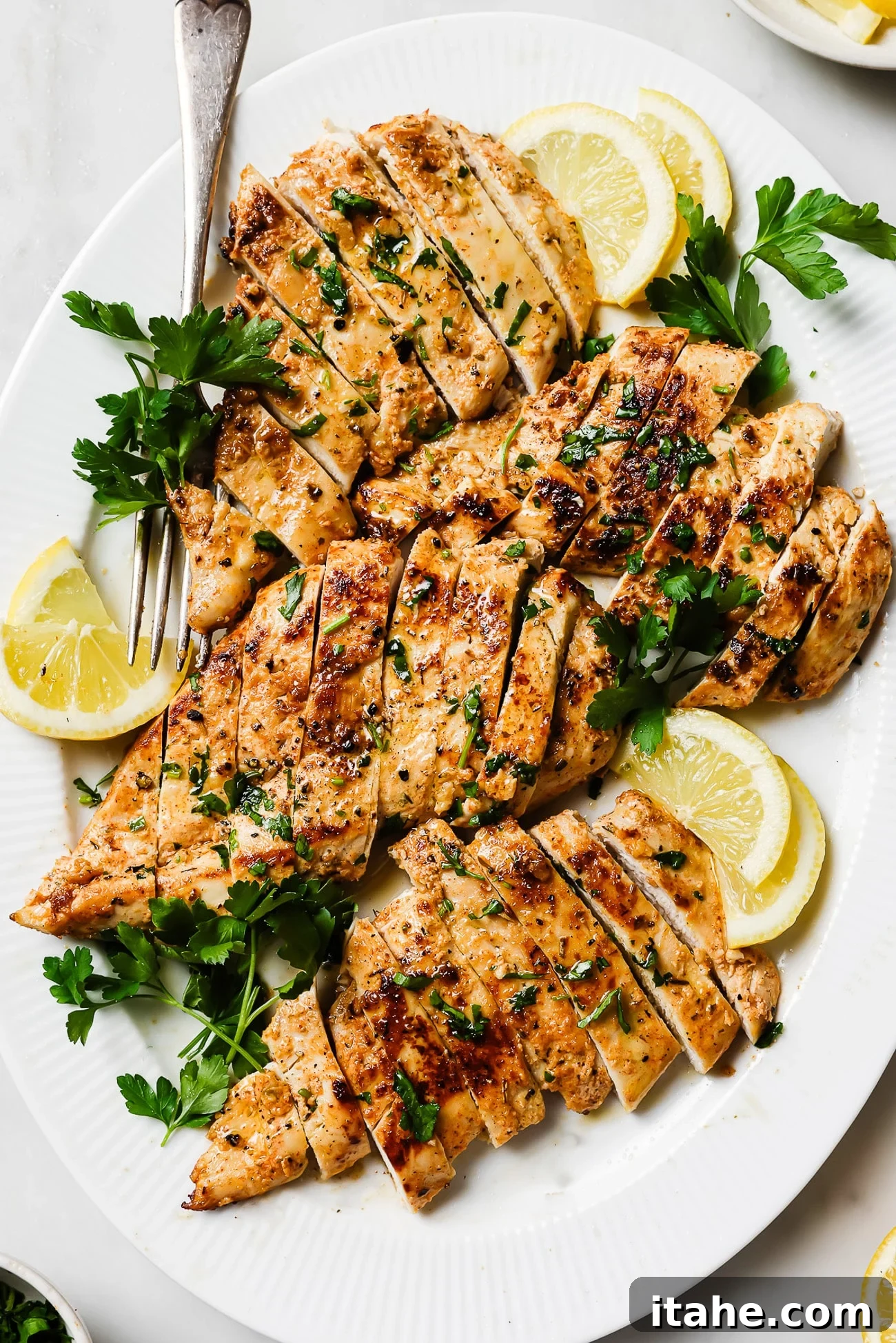 Creamy Yogurt Chicken Breasts 2 Yogurt-marinated chicken breast sliced and garnished with parsley and lemon wedges on a white serving plate.