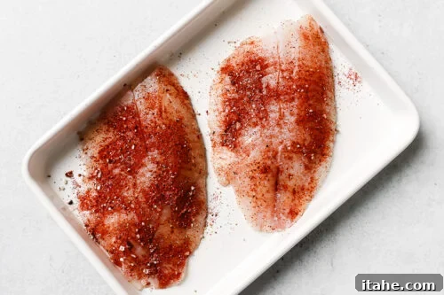 Seasoned raw tilapia fillet on a white baking sheet.
