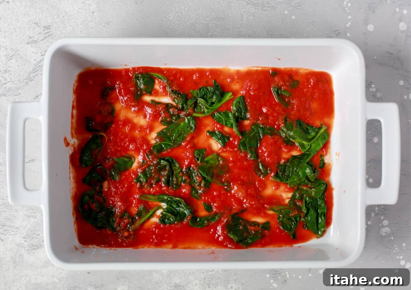 Noodle-Free Zucchini Lasagna 3 Overhead view of a white casserole dish with a layer of vibrant red tomato sauce and green spinach.