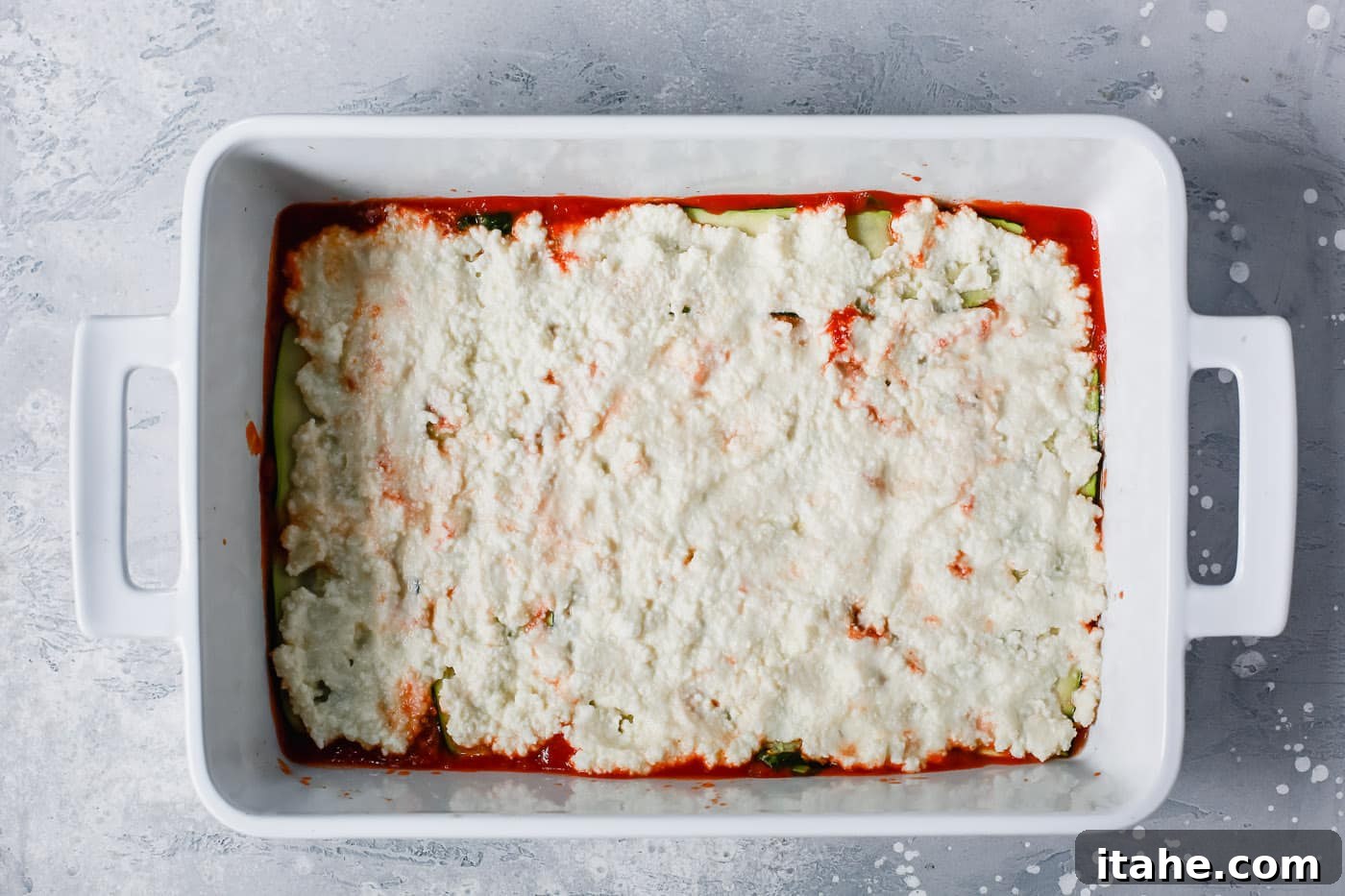 Noodle-Free Zucchini Lasagna 5 Overhead view of a white casserole dish with a layer of creamy white ricotta cheese spread over the zucchini.