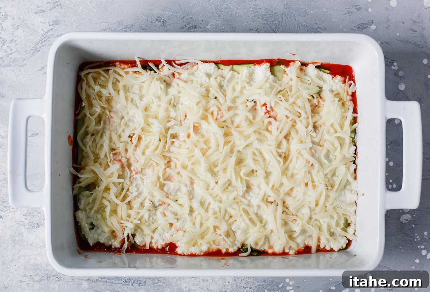 Noodle-Free Zucchini Lasagna 6 Overhead view of a white casserole dish showing shredded mozzarella cheese covering the ricotta and zucchini layers.