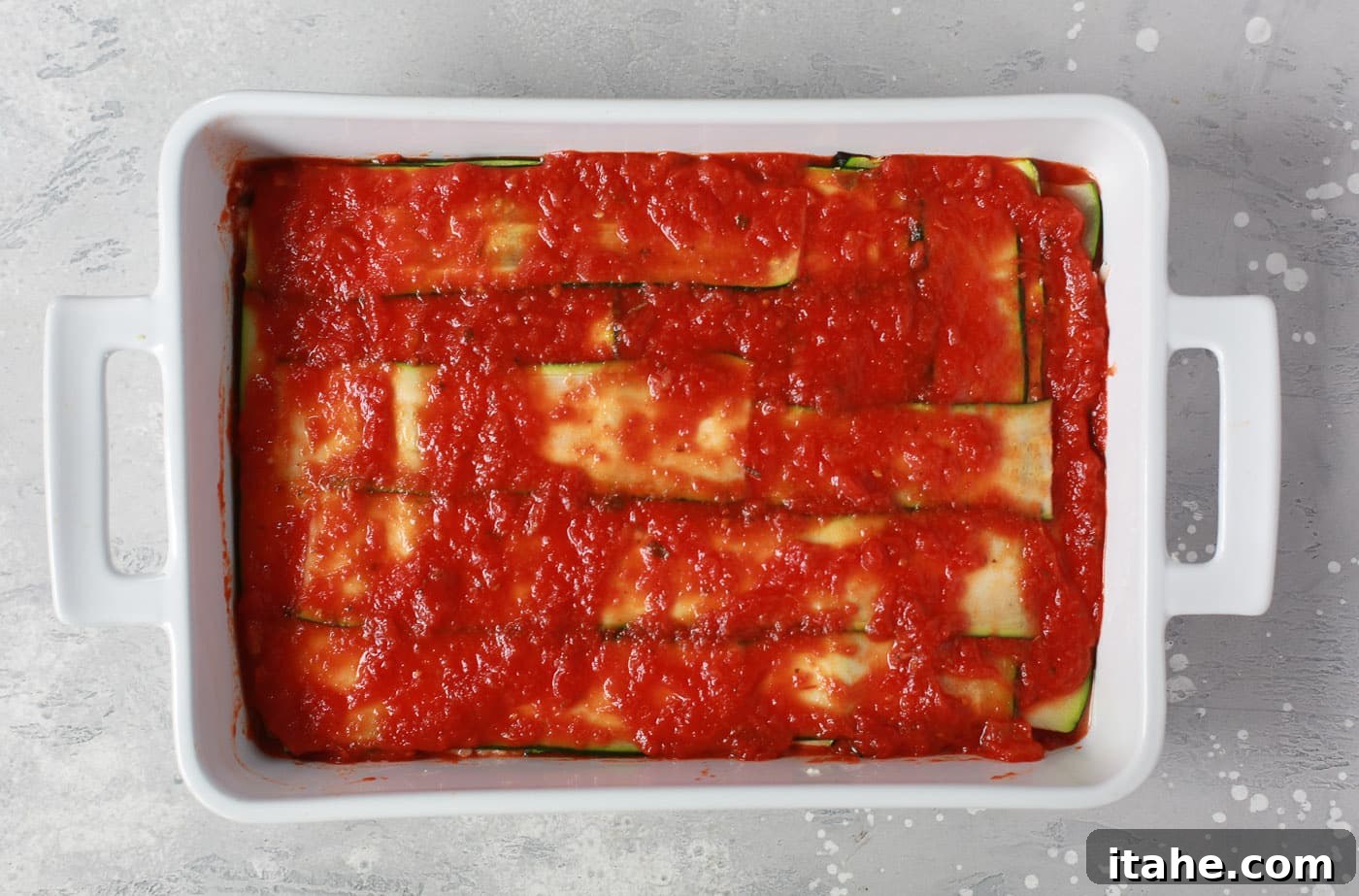 Noodle-Free Zucchini Lasagna 7 Overhead view of a white casserole dish showing the repeated layers of zucchini and tomato sauce during assembly.