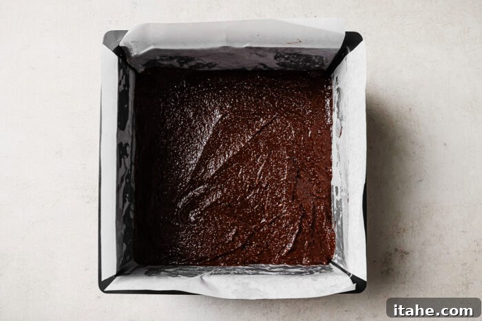 Chocolate brownie batter evenly spread in a parchment-lined square baking pan, ready to bake.