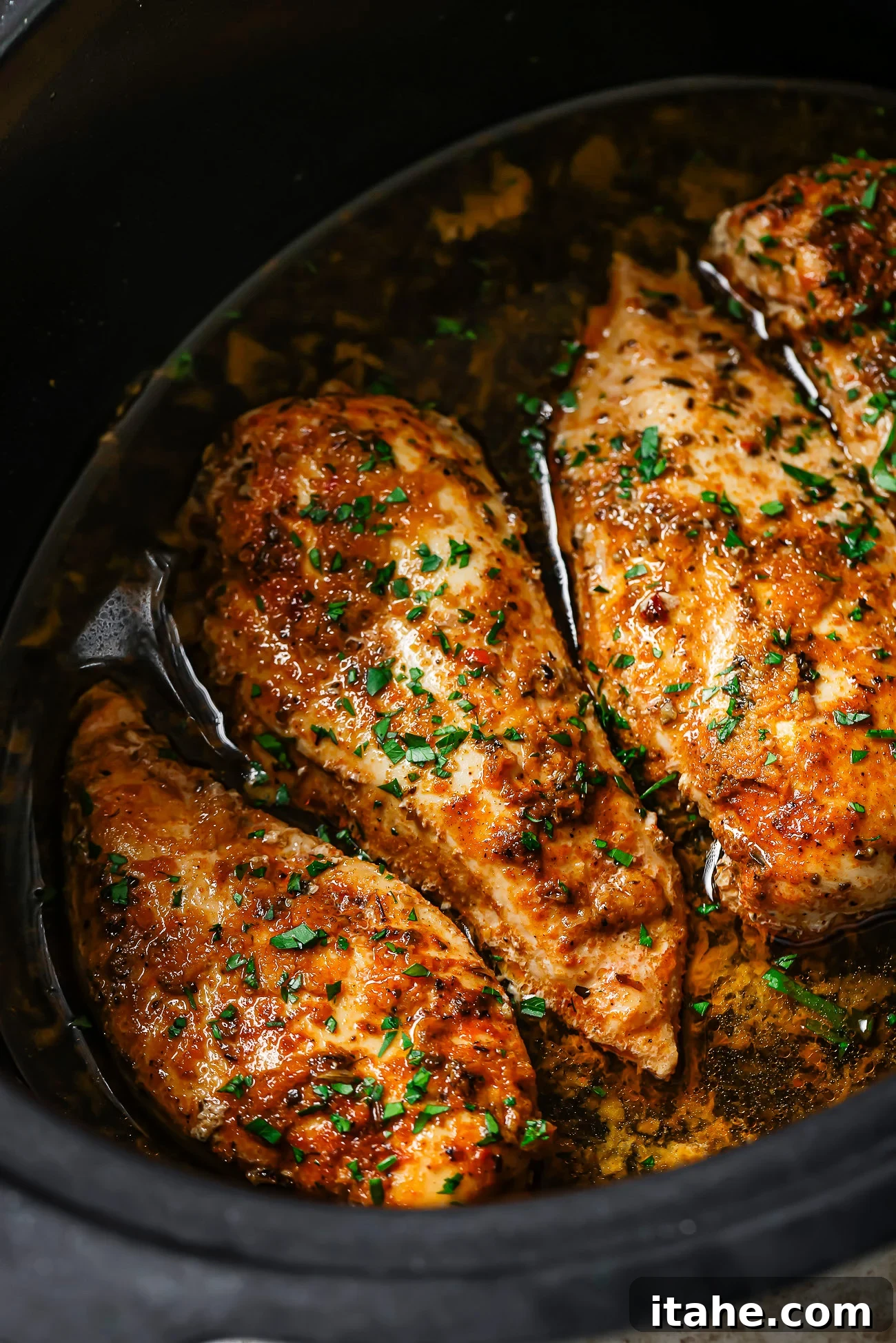Three seasoned Slow Cooker Chicken Breasts sit in a dark slow cooker with flavorful sauce and sprinkled fresh herbs, showcasing a juicy and tender appearance.