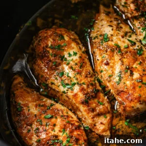 Three seasoned Slow Cooker Chicken Breasts sit in a dark slow cooker with flavorful sauce and sprinkled fresh herbs, showcasing a juicy and tender appearance.