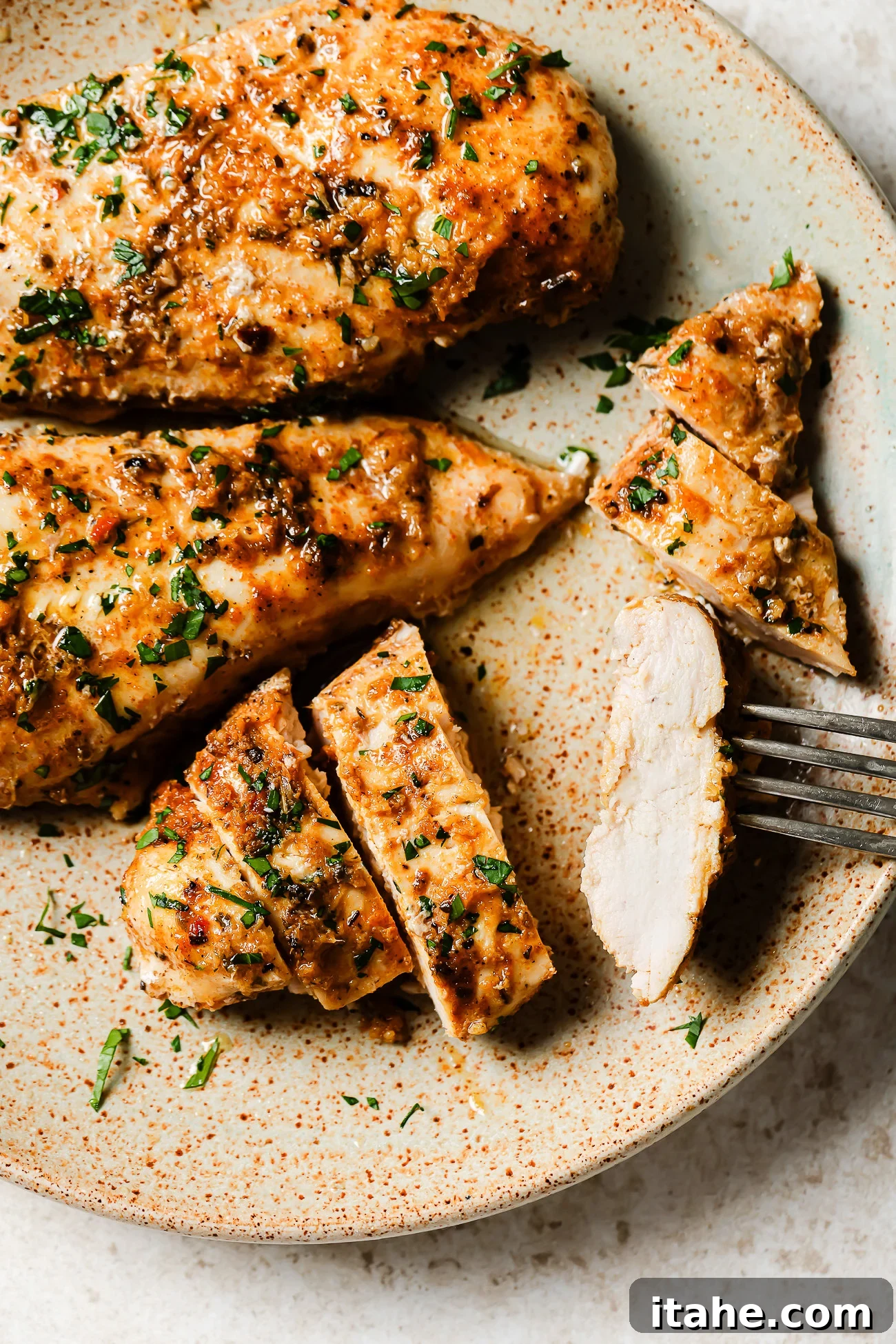 A plate with cooked, seasoned Slow Cooker Chicken Breast garnished with chopped herbs. One chicken breast is sliced, and a fork is holding a piece. The food is presented on a rustic, speckled plate.