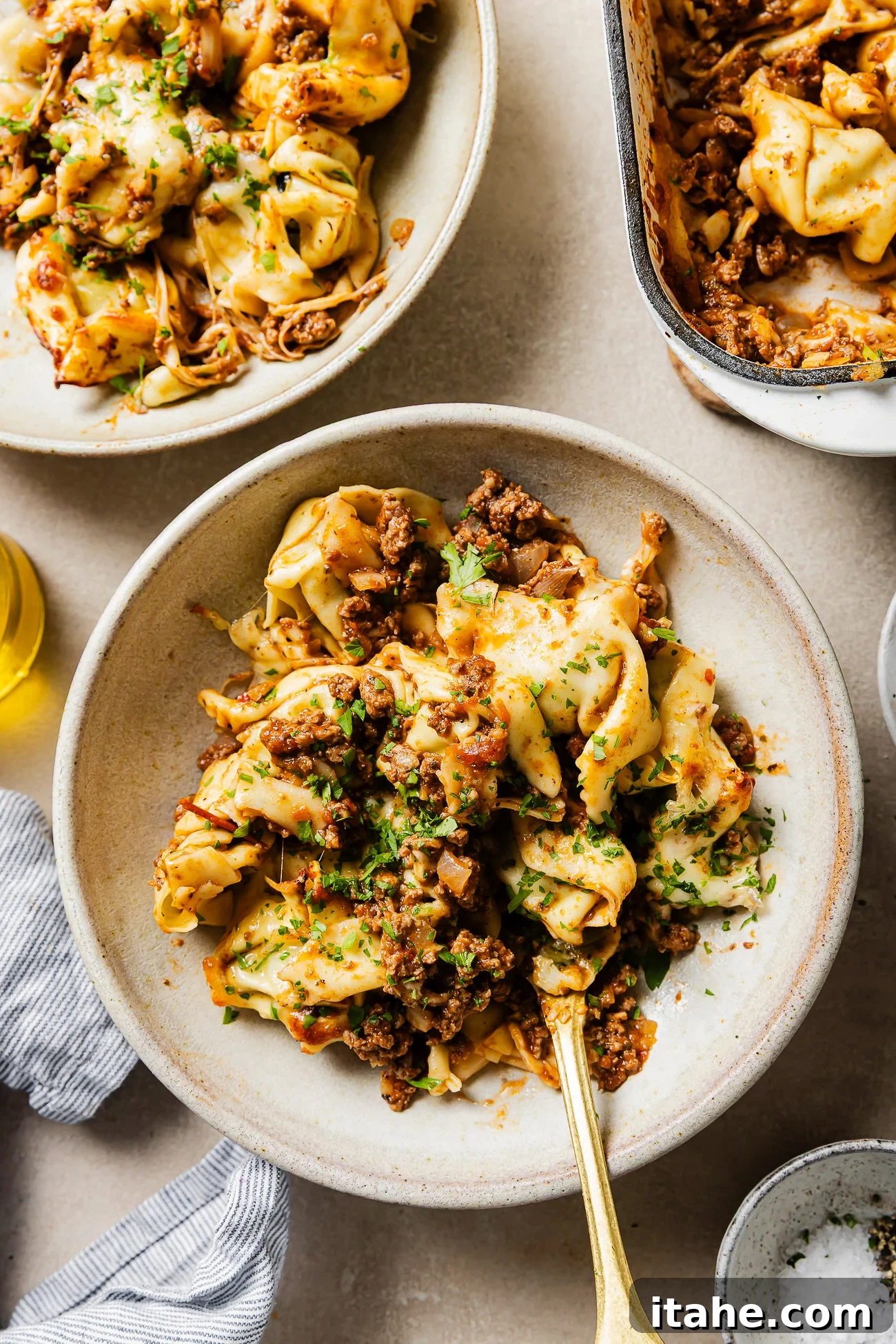 Cheesy Baked Tortellini with Seasoned Ground Beef 10 A bowl of tortellini pasta topped with a hearty meat sauce and fresh herbs, with a fork in the bowl. There are more servings of pasta in dishes nearby on a light-colored surface.