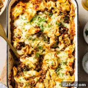 Cheesy Baked Tortellini with Seasoned Ground Beef 11 A baked pasta casserole in a white dish filled with cheese, ground meat, and herbs, topped with melted cheese and chopped parsley. A serving spoon is placed in the dish, ready for serving.