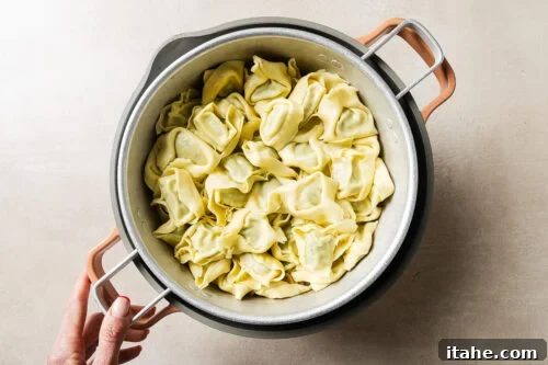Cheesy Baked Tortellini with Seasoned Ground Beef 12 A hand holds a pot filled with uncooked tortellini pasta on a light-colored surface.