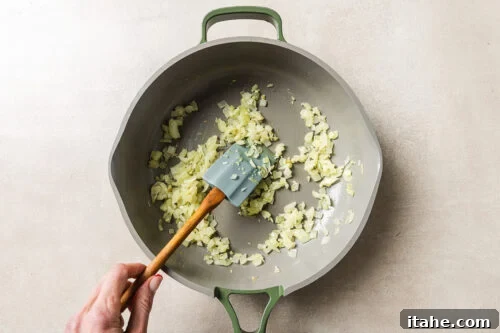 Cheesy Baked Tortellini with Seasoned Ground Beef 13 A hand stirs chopped onions in a large, light-colored pan with a green handle, using a spatula. The onions are being sautéed on a light-colored surface.
