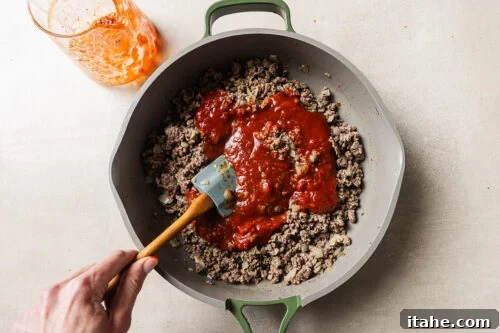 Cheesy Baked Tortellini with Seasoned Ground Beef 14 A hand stirs browned ground beef and chopped onions in a skillet with tomato sauce being mixed in. A glass with leftover sauce sits nearby on a light surface.