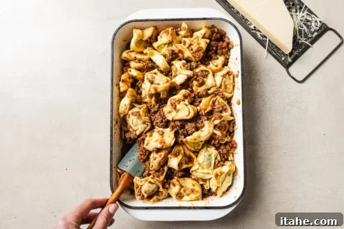 Cheesy Baked Tortellini with Seasoned Ground Beef 15 A hand holds a spatula in a baking dish filled with tortellini pasta and meat sauce. A block of Parmesan cheese and a grater sit on the surface nearby.