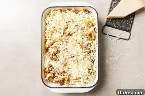 Cheesy Baked Tortellini with Seasoned Ground Beef 16 A rectangular baking dish filled with uncooked pasta topped with shredded cheese sits on a light surface. Next to it are a metal grater and a wedge of cheese, with cheese shavings scattered around.