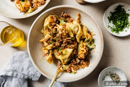 Cheesy Baked Tortellini with Seasoned Ground Beef 17 A bowl of pasta with crumbled ground meat, garnished with chopped herbs. Surrounding the bowl are a dish of salt and pepper, a small bowl of herbs, a glass container of olive oil, and a striped napkin.