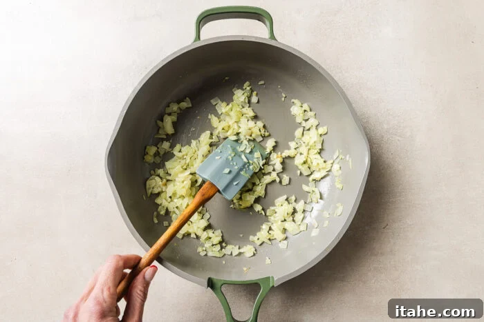Cheesy Baked Tortellini with Seasoned Ground Beef 5 A hand stirs chopped onions in a large, light-colored pan with a green handle, using a spatula. The onions are being sautéed on a light-colored surface.