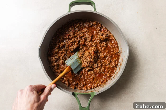 Cheesy Baked Tortellini with Seasoned Ground Beef 7 A hand stirs seasoned ground beef and tomato sauce with a green spatula in a large gray pot on a light countertop.