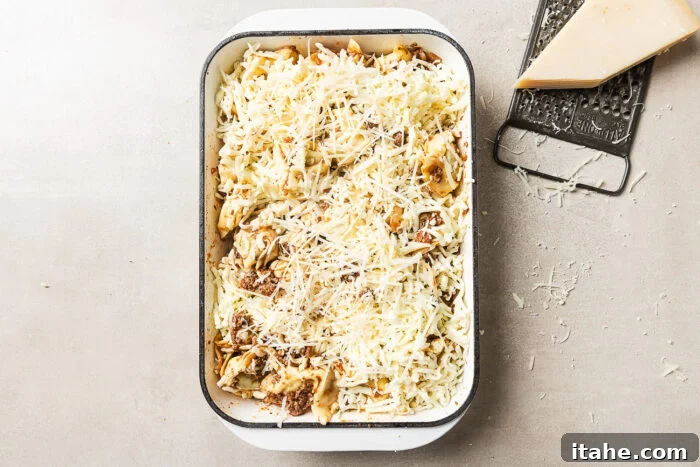 Cheesy Baked Tortellini with Seasoned Ground Beef 9 A rectangular baking dish filled with uncooked pasta topped with shredded cheese sits on a light surface. Next to it are a metal grater and a wedge of cheese, with cheese shavings scattered around.