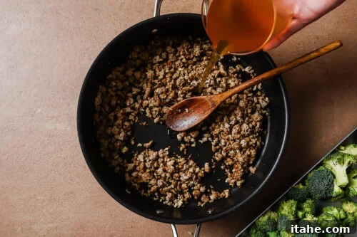 A hand pours broth from a measuring cup into a skillet with browned ground meat, stirred by a wooden spoon. Fresh broccoli is visible on a tray beside the skillet.