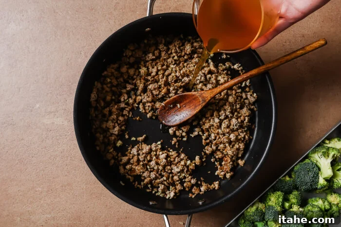 A hand pours broth from a measuring cup into a skillet with browned ground meat, stirred by a wooden spoon. Fresh broccoli is visible on a tray beside the skillet.