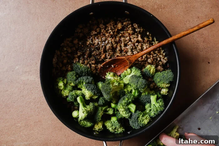 A black pan on a brown surface contains cooked ground meat and fresh broccoli florets being stirred with a wooden spoon. A hand is adding more broccoli to the pan from a metal tray.