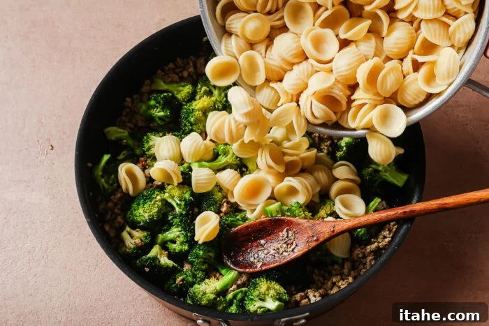 Cooked orecchiette pasta is being poured from a pot into a skillet containing sautéed broccoli and ground meat, with a wooden spoon resting in the mixture.