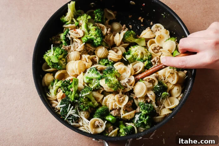 A hand stirs orecchiette pasta with broccoli, mushrooms, grated cheese, and ground meat in a black skillet on a light brown surface.