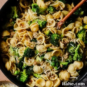 A skillet filled with orecchiette pasta, broccoli florets, crumbled sausage, and grated Parmesan cheese, with a wooden spoon and a bowl of extra cheese nearby on a striped cloth.