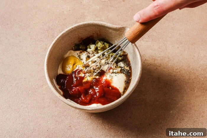 A hand uses a whisk to mix ingredients including ketchup, mayonnaise, mustard, relish, and seasonings in a small beige bowl on a tan countertop.